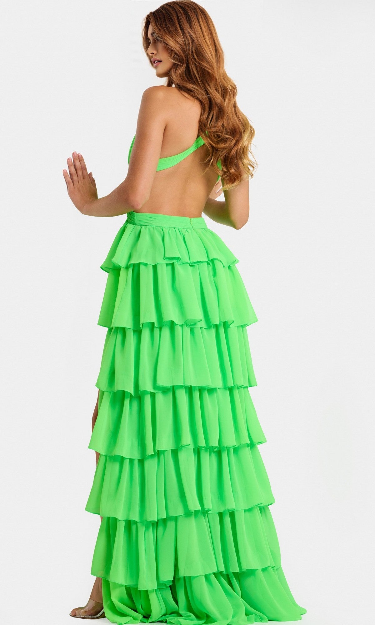 A woman with long wavy hair poses against a white background wearing the Jovani 46338, a bright green halter chiffon long prom dress with an open back and tiered ruffle skirt.